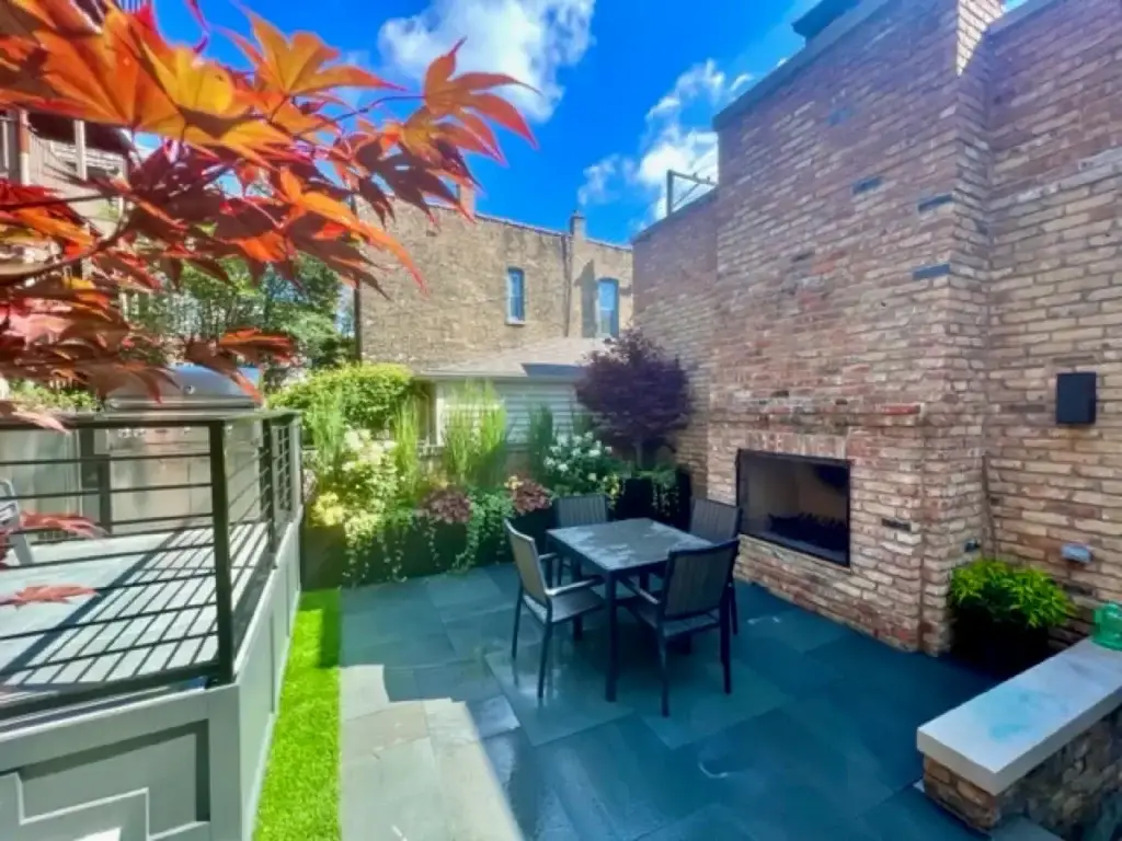 Modern Chicago patio with dark stone pavers, built-in brick fireplace, contemporary dining set, integrated greenery, and layered landscaping in a compact urban backyard.