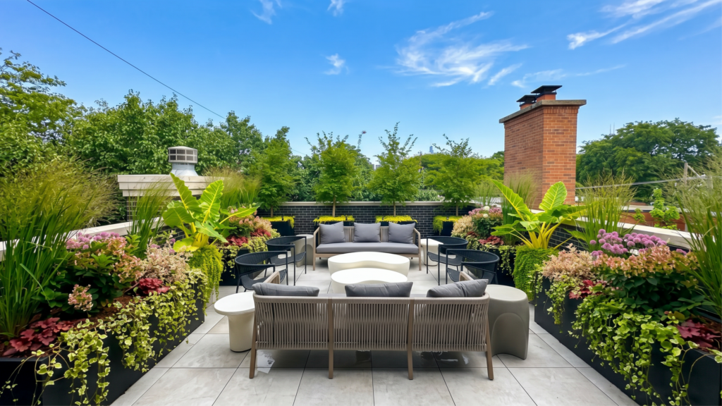 A luxury Chicago rooftop terrace with large custom black flower boxes filled with tropical plants, ornamental grasses, and colorful seasonal flowers surrounding a modern outdoor seating area under a blue sky. @keyframes bannerSlideUp { from { opacity: 0; transform: translateY(8px); } to { opacity: 1; transform: translateY(0); } }