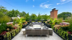 A luxury Chicago rooftop terrace with large custom black flower boxes filled with tropical plants, ornamental grasses, and colorful seasonal flowers surrounding a modern outdoor seating area under a blue sky. @keyframes bannerSlideUp { from { opacity: 0; transform: translateY(8px); } to { opacity: 1; transform: translateY(0); } }