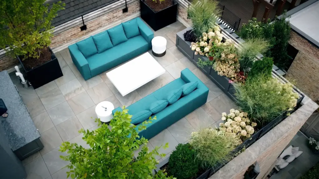 Aerial view of a Chicago rooftop deck with two teal sofas facing each other across a white coffee table on gray porcelain pavers, framed by black planters with small trees, built-in planter boxes filled with hydrangeas and ornamental grasses, and an outdoor kitchen counter along the brick wall.