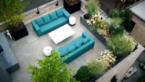 Aerial view of a Chicago rooftop deck with two teal sofas facing each other across a white coffee table on gray porcelain pavers, framed by black planters with small trees, built-in planter boxes filled with hydrangeas and ornamental grasses, and an outdoor kitchen counter along the brick wall.
