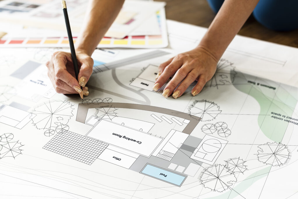 A designer's hands hold a pencil over a detailed landscape architectural floor plan showing outdoor spaces, a pool, and plantings, with color palette swatches in the background, representing the design-build planning process for a luxury outdoor project.