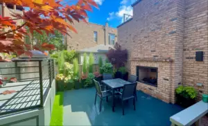 A city backyard with a blue-gray paver patio, dark metal dining set, built-in brick fireplace, tall layered privacy plantings along the fence line, and a stainless steel grill on an adjacent deck, framed by red Japanese maple leaves in the foreground.