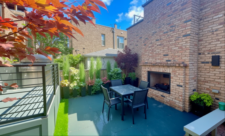 A city backyard with a blue-gray paver patio, dark metal dining set, built-in brick fireplace, tall layered privacy plantings along the fence line, and a stainless steel grill on an adjacent deck, framed by red Japanese maple leaves in the foreground.