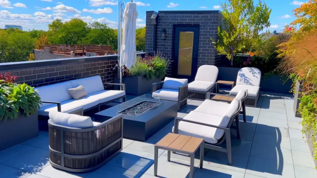 A rooftop deck with gray tile flooring, a rectangular fire pit surrounded by a sofa and lounge chairs with white cushions, a wicker armchair, potted plants and ornamental grasses along the perimeter, and a dark brick structure with a glass-paneled door, framed by trees and a city skyline.